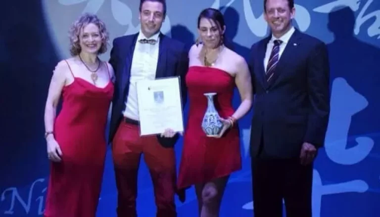 Four people are standing together, smiling at the camera. Two women in red dresses are on either end, while two men in formal attire are in the middle. One man is holding a certificate, and the woman next to him is holding a vase. They appear to be at an event.