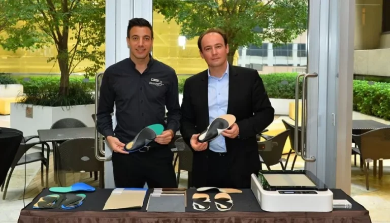 Two men stand behind a table displaying various orthotic insoles and related materials. Both are dressed in business attire, one in a black shirt and the other in a suit, each holding insoles. In the background, there are large windows showing a garden-like outdoor area.