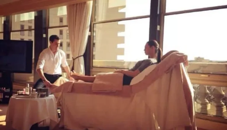 A man is giving a foot massage to a woman who is lying on a lounge chair in a sunlit room with large windows. The room has a calm, relaxing ambiance, with city buildings visible outside the windows. A table with massage supplies is beside them.