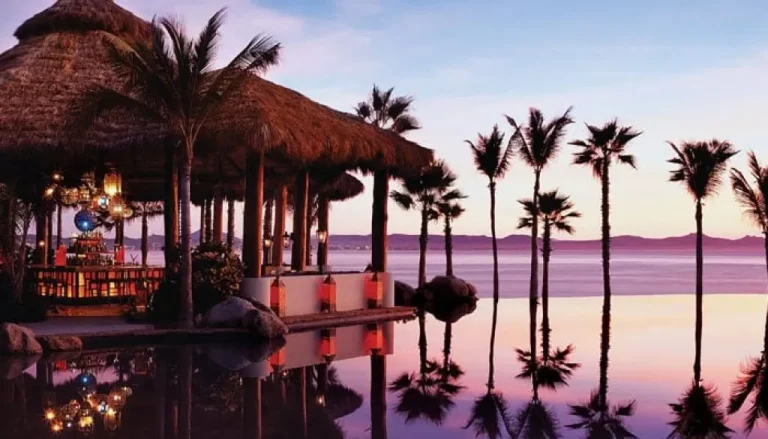 A tropical bar with a thatched roof stands beside a serene infinity pool. The pool reflects tall palm trees and a colorful sunset sky. Tables and hanging lanterns are visible inside the bar, creating a cozy and inviting atmosphere.