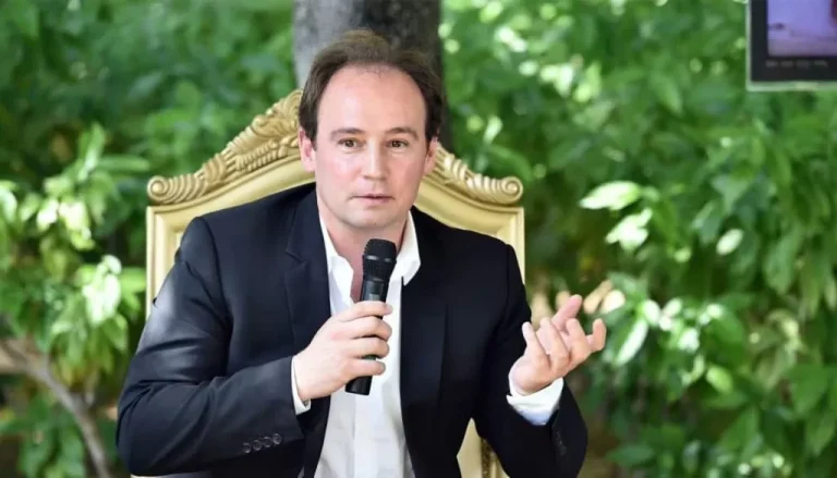 Un homme en costume noir et chemise blanche est assis à l’extérieur sur une chaise d’apparence royale, tenant un microphone dans une main et faisant des gestes de l’autre. Il semble parler, avec un fond vert et luxuriant d'arbres et de feuillages derrière lui.