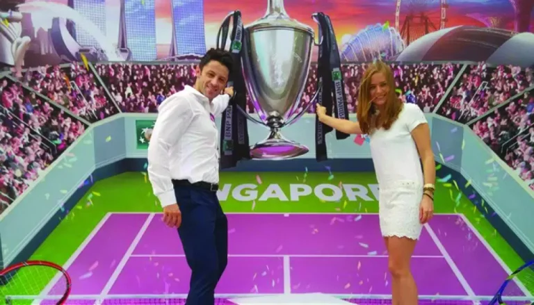 A man in a white shirt and blue pants, and a woman in a white dress are standing on a purple tennis court with a backdrop of a cheering crowd and cityscape, both holding a large silver trophy. The word "Singapore" is visible on the court surface.