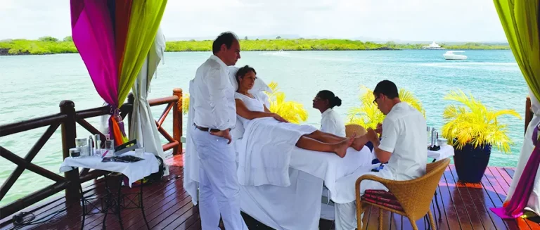 Two people in white outfits provide a spa treatment to a person lying on a massage table under a canopy by the water. The scene overlooks a tranquil blue sea with lush green vegetation in the background. The setup is on a wooden deck with vibrant drapes.