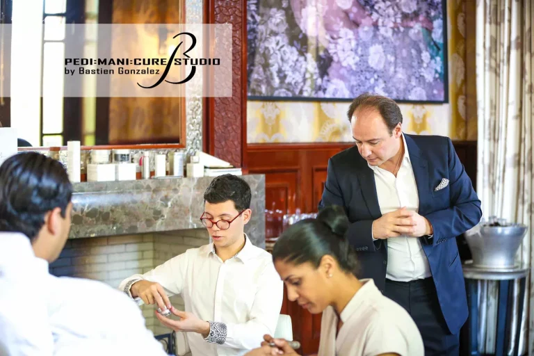 A man in a suit observes while three people, two men and one woman, focus on a task at a table in a well-lit room. The wall has elegant decor and a large mirror. A logo in the top-left corner reads "PEDI:MANI:CURE STUDIO by Bastien Gonzalez.