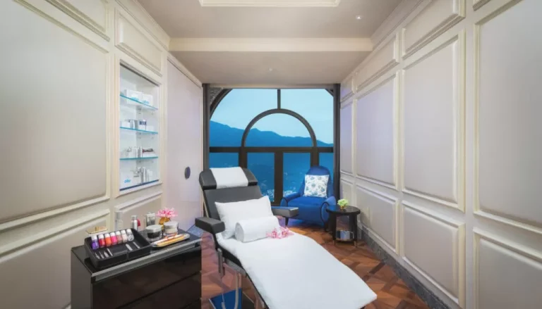 A serene spa room featuring a reclining chair with white towels, a table with an array of nail polishes, and a side table holding a cup and essential oils. A large arched window offers a mountain view, while soft white walls and wooden floors create a calm atmosphere.