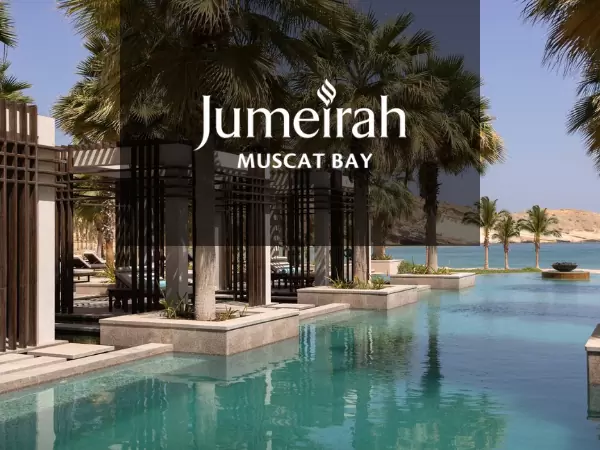 Image of a luxurious poolside at Jumeirah Muscat Bay featuring clear blue water, palm trees, and modern architectural elements. The shoreline and mountains are visible in the background under a bright blue sky. The resort's name is prominently displayed.