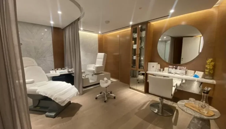 A modern spa treatment room featuring two reclining chairs with white fabric covers, a round mirror above a counter with skincare products, a soft curtain dividing the space, and a wooden floor with warm lighting.