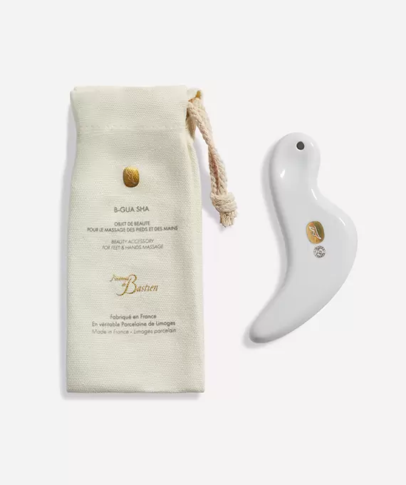 A white, smooth, bird-shaped massage tool is placed next to a beige drawstring pouch. The pouch has product information printed on it in both French and English, with a gold logo above the text. The massage tool also features a small gold logo.