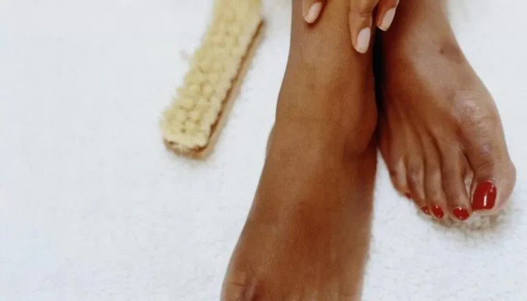 Gros plan d'une paire de pieds sur une serviette blanche. Un pied repose sur l’autre. Les ongles sont polis en rouge et une main touche doucement le dessus du pied. Une brosse à pieds avec un manche et des poils en bois se trouve en arrière-plan.