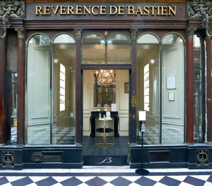 Une devanture soignée avec l'enseigne "Révérence de Bastien" au-dessus de grandes fenêtres cintrées et d'une porte vitrée. À l’intérieur, un lustre est suspendu au-dessus d’une élégante table noire. L'entrée présente d'élégantes colonnes et un sol en damier, suggérant un cadre luxueux.