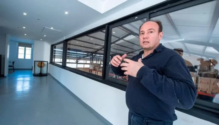 Un homme vêtu d'une chemise bleu marine fait des gestes avec ses mains alors qu'il se tient dans un couloir avec de grandes fenêtres. Derrière les fenêtres, il semble y avoir un espace industriel ou un entrepôt avec des étagères et des cartons. Le couloir est bien éclairé par des plafonniers.