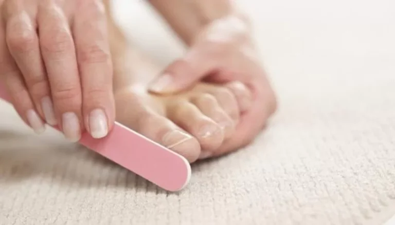 Une personne se lime les ongles des pieds avec une lime à ongles rose, en se concentrant sur ses pieds nus. Les mains et les pieds de la personne sont au centre de l'attention, avec une surface beige et texturée en dessous. L’image représente probablement une activité de soin des pieds ou de toilettage.