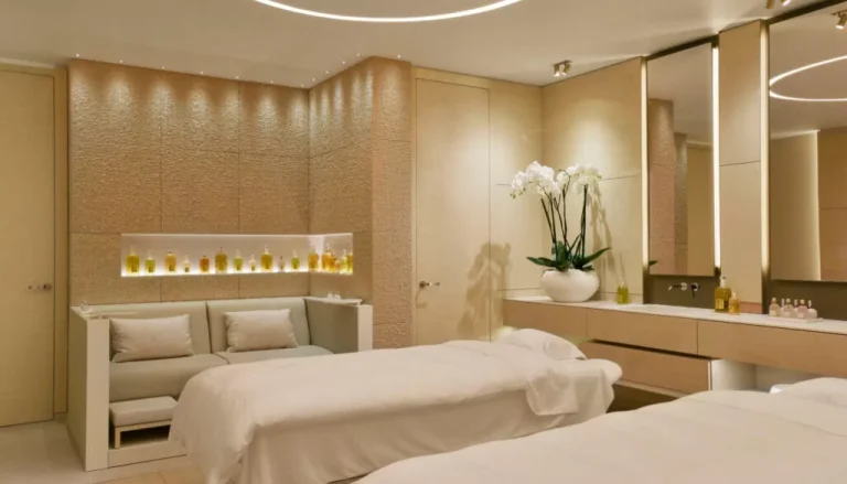 A luxurious spa room with soft lighting features two massage tables with white linens, a comfortable seating area, and a variety of neatly arranged essential oils on shelves. Elegant decor elements include a large vase with white orchids on a sleek counter.