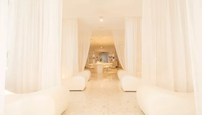 A serene, minimalist lounge area with white draped curtains and sleek white seating. The space is bathed in soft light, enhancing the clean and tranquil atmosphere. A table is centered in the background, creating an inviting focal point.