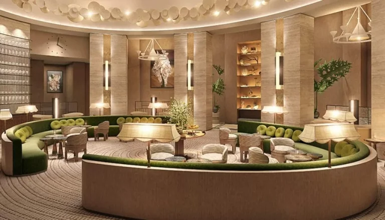 A luxurious hotel lobby with elegant, curved seating areas arranged around a central table adorned with greenery. The space features soft lighting, tall columns, modern decor, and an array of comfortable chairs and tables. Warm, neutral tones dominate the decor.