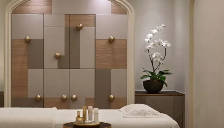 A modern spa room featuring a massage table with a white towel and various small bottles on a round side table. The background has a stylish, geometric wall panel design with a potted orchid plant placed on a cabinet. The room has a calm, neutral color palette.