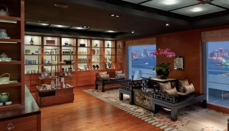 A luxurious, modern living room with a wooden floor and ceiling. It features elegant furniture, including a sofa, armchairs, and a coffee table. A large display shelf with various items is on the left. Large windows reveal a city skyline and waterfront view.