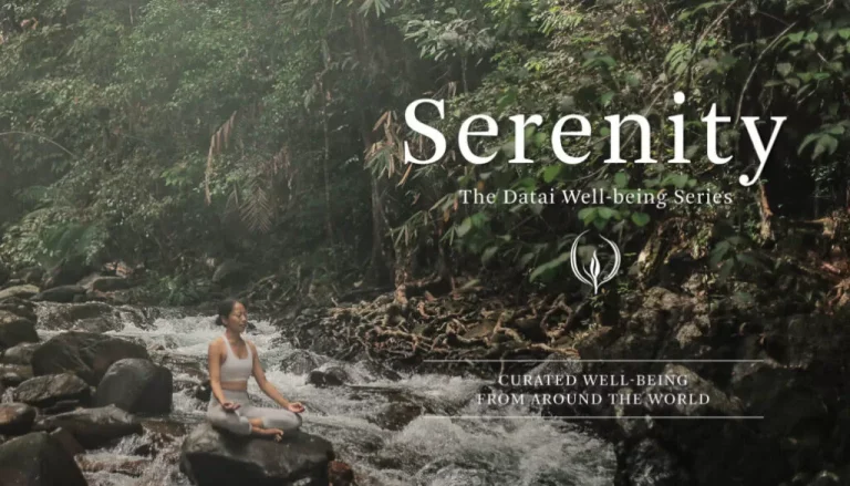 A person meditates on a large rock in a stream surrounded by dense greenery. Text on the image reads: "Serenity - The Datai Well-being Series - Curated well-being from around the world.