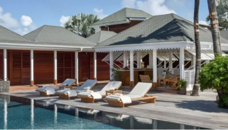 Un luxueux complexe tropical dispose de plusieurs chaises longues blanches au bord d’une piscine étincelante. Derrière la piscine, il y a une terrasse en bois avec un coin salon ombragé, des palmiers et des bâtiments aux toits à pignon verts. Le ciel est bleu avec quelques nuages épars.