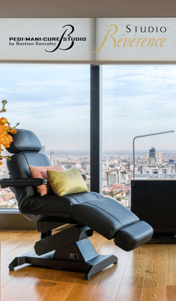 Un studio de manucure et pédicure moderne avec un fauteuil inclinable placé à côté d'une grande fenêtre offrant une vue sur un paysage urbain. Des éléments décoratifs, comme un vase à fleurs orange, ajoutent à l'élégance. Le texte « Studio Reverence » s'affiche en haut.