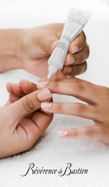 Une main tenant un petit tube applique une goutte de produit sur l'ongle d'une autre personne. Les mains reposent sur une surface blanche. Sous les aiguilles, le texte "Révérence de Bastien" est visible.