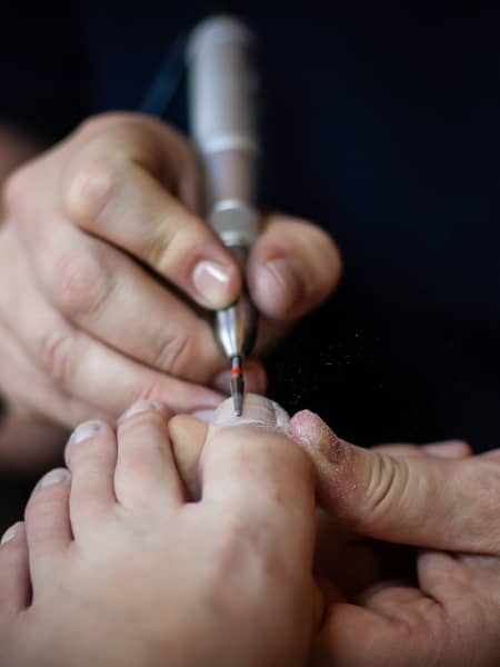 Close-up of a person receiving a pedicure. Another individual is using a nail drill to shape the toenails. The focused area shows the person's toes and the hands of the person performing the pedicure, with fine nail dust visible in the air.